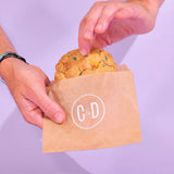 Birthday Cake New York Cookie by Crumbs & Doilies in brown wrapper with hands holding the cookie against a pale purple background